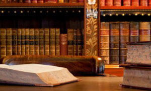 Aged criminal law book open on solid wood library table with more leather bound books lining the shelves behind the table.